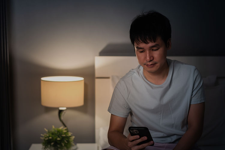 薄暗い寝室でスマートフォンを見ているTシャツの男性