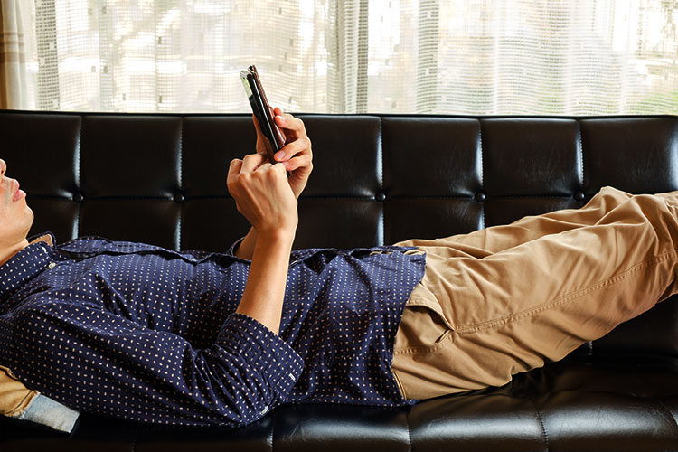 ソファの上に寝そべってスマホを操作する男性の写真